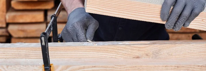Person verklebt Holzbalken, die mit einer Schraubzwinge zusammengehalten werden