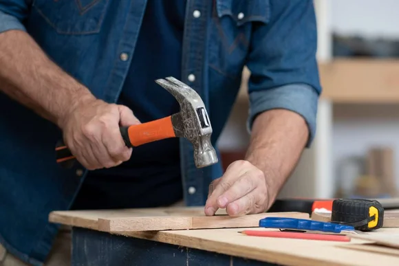 Eine Person schlägt mit einem Hammer einen Nagel in ein Holzbrett.