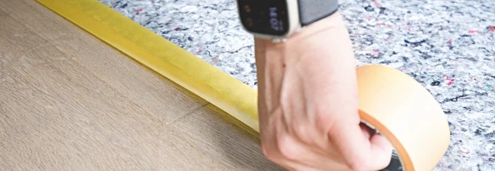 Person klebt Malerkrepp auf einen Holzboden