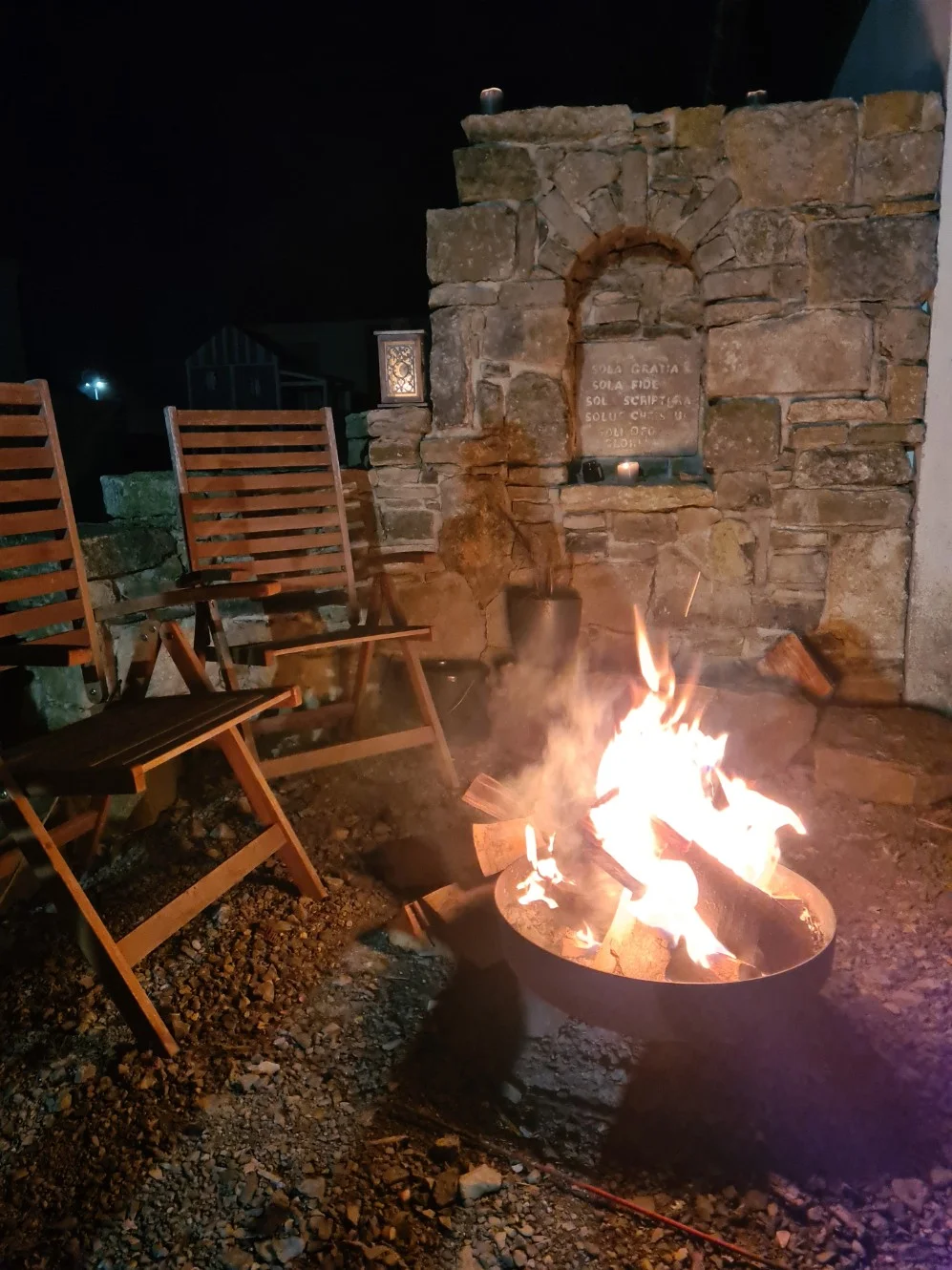 Zwei Holzstühle an einer brennenden Feuerschale vor einer Steinmauer bei Nacht.