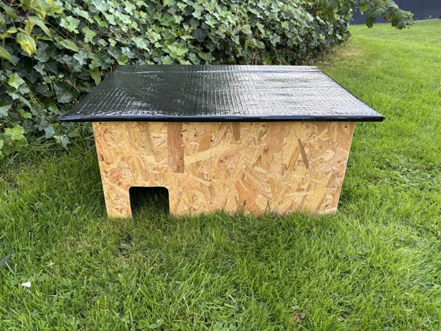 Ein Igelhaus aus Holzplatten mit schwarzem Dach steht auf einer Wiese.