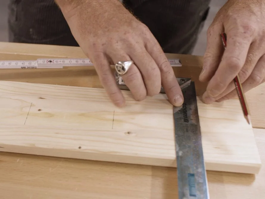 Zwei Hände richten einen Winkel auf einem Holzbrett mit einem Schreinerwinkel und einem Bleistift aus.