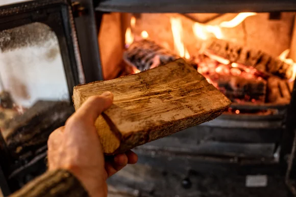 Ein Holzscheit wird in einen brennenden Kaminofen gelegt.