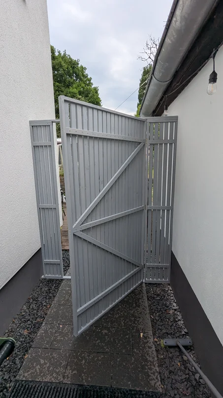 Ein offenes, graues Gartentor in einem Durchgang zwischen zwei Hauswänden.