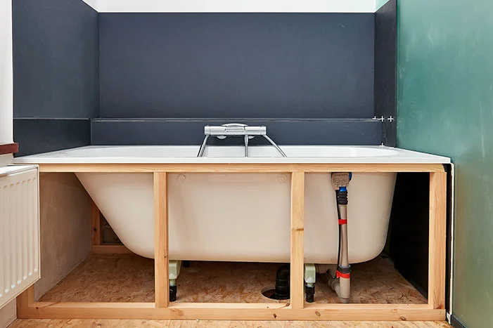 Badewanne mit sichtbarem Abfluss und Wasserleitungen auf Holzkonstruktion.