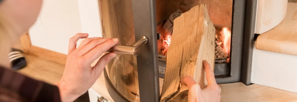 Eine Person legt ein Stück Holz in einen brennenden Kaminofen.