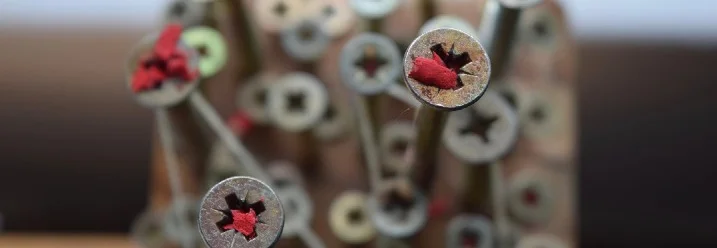 Schrauben in einem Holzbrett und teilweise stecken rote Stoffreste in den Schraubenköpfen