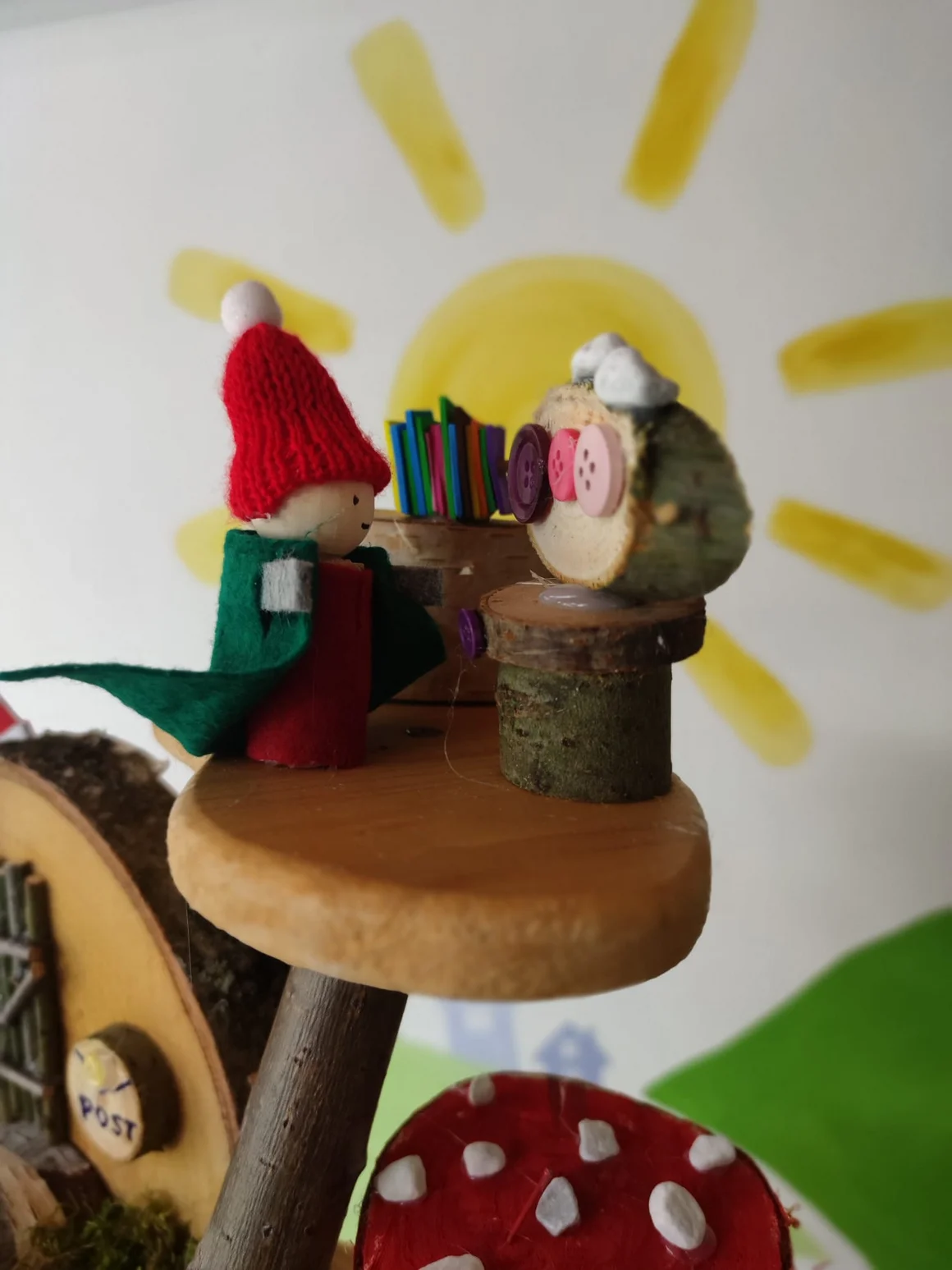 Zwei gebastelte Holzfiguren, ein Wichtel und eine Eule, vor einer gemalten Sonne.