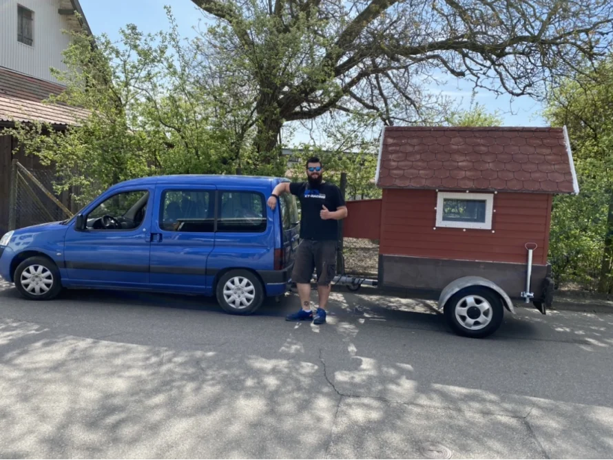 Person steht neben einem roten Mini-Holzhaus auf Rädern