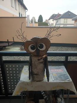 Eine aus Holz gebastelte Eule mit großen Augen steht auf einem Balkontisch.